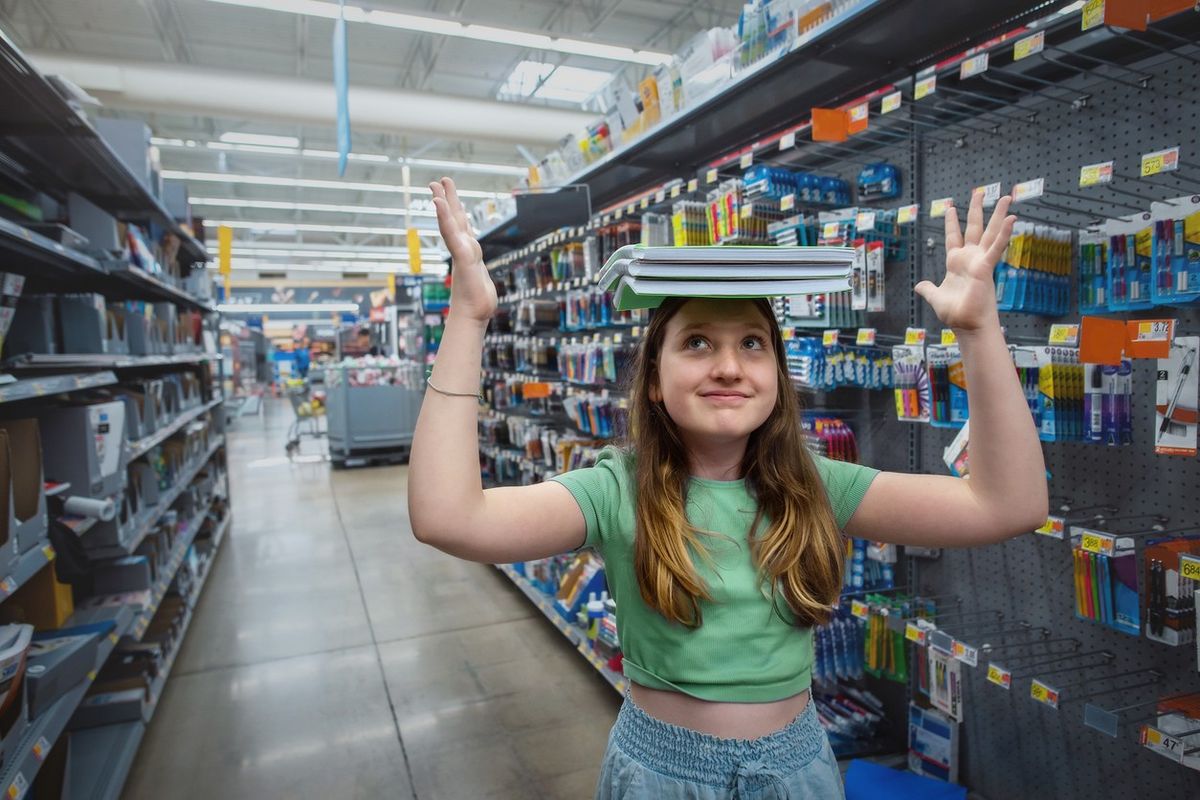 Girl balancing school supplies on head as she shops for notebooks in store aisle