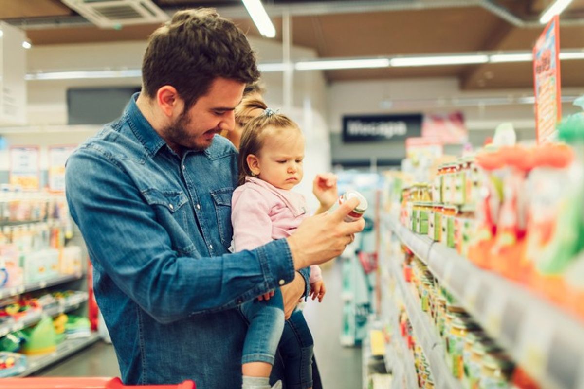 Person in denim shirt with child in pink jacket examines baby food pouch in store aisle. Shelves hold brands like Ella’s Kitchen, Heinz.