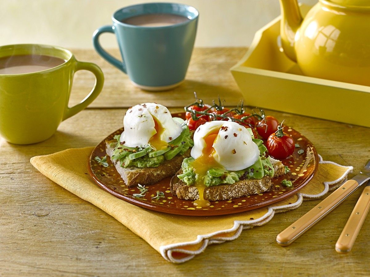 Poached eggs on avocado toast with tomatoes and mugs of tea on table
