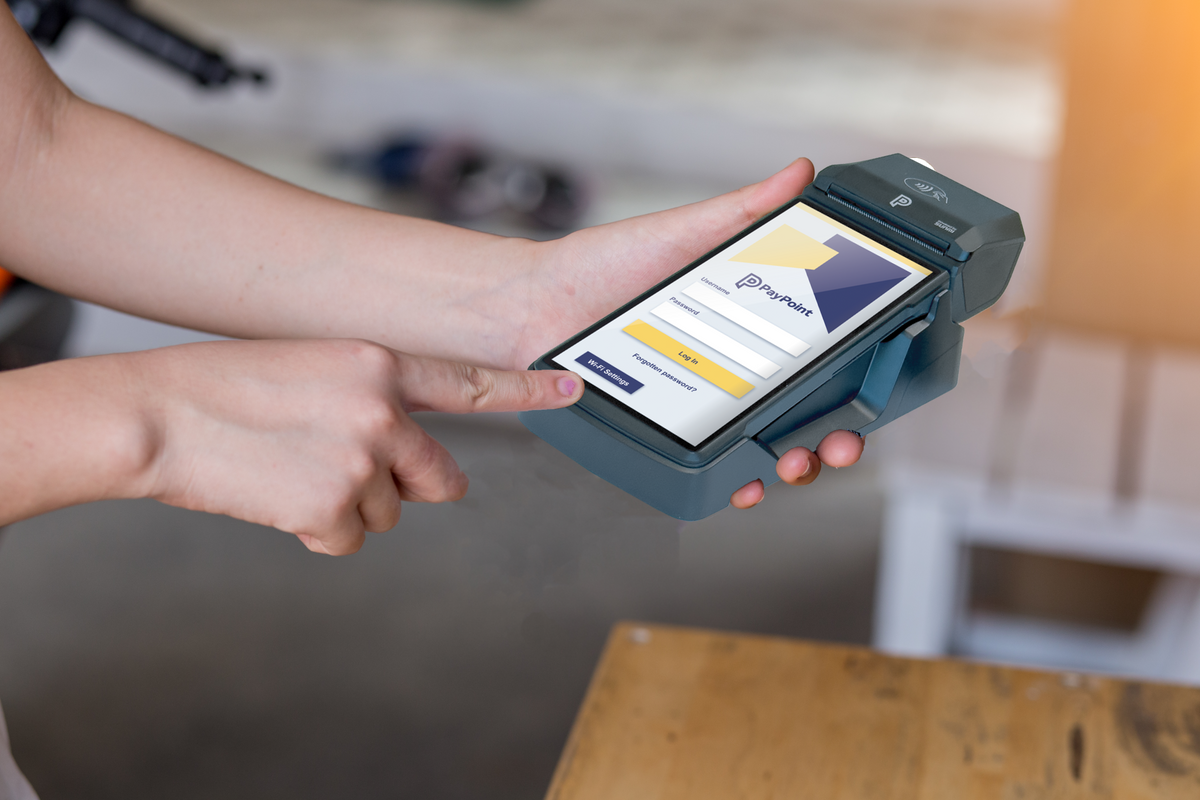 Retailer using PayPoint handheld device showing login screen for mobile payment services.