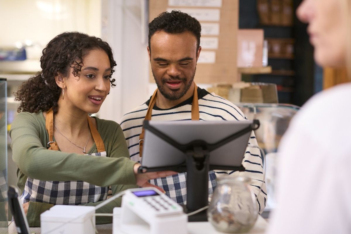 Woman Working In Food Shop Training Man With Down Syndrome How To Use Checkout